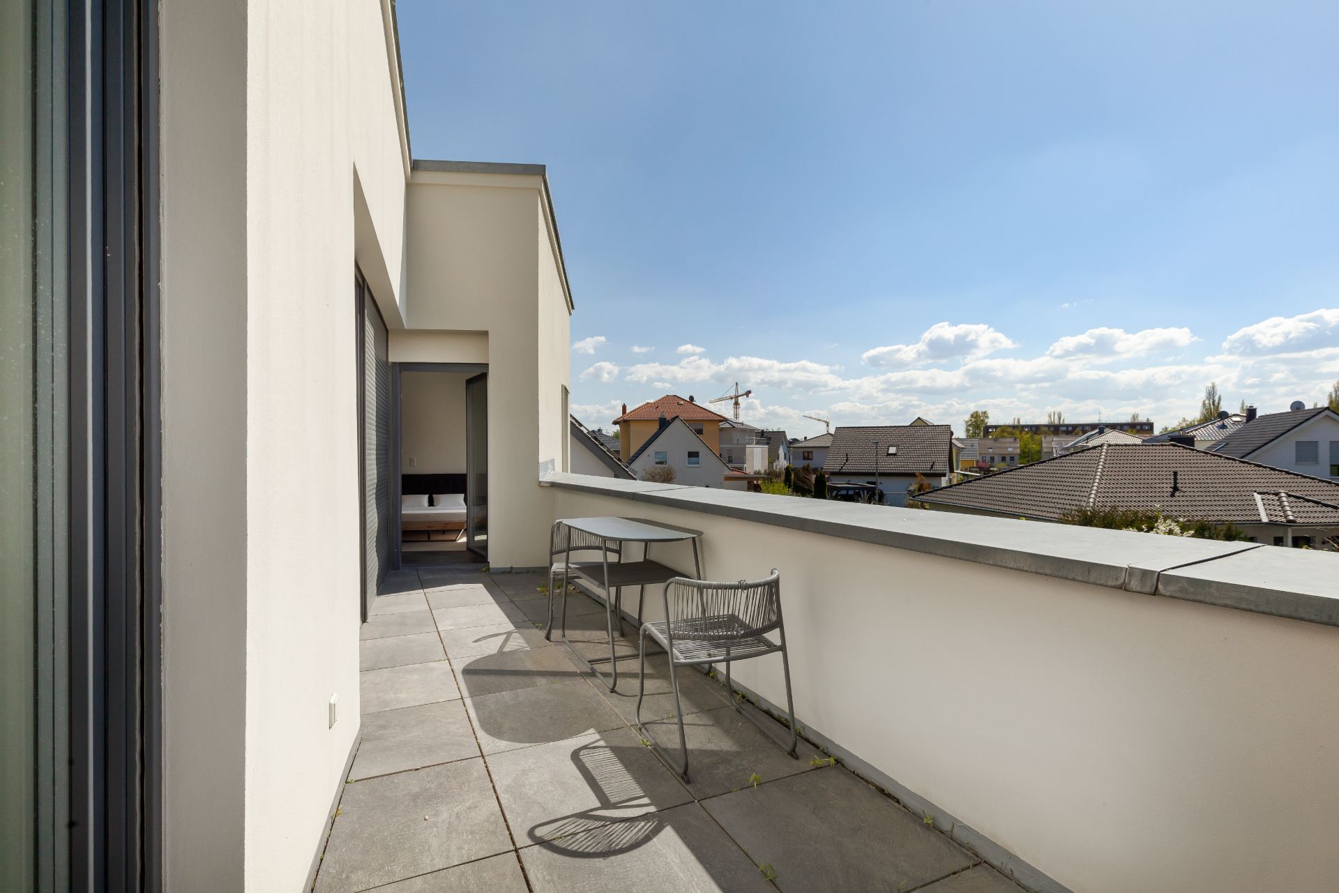 Geräumiger Balkon mit zwei Stühlen und einem Tisch, Blick auf Nachbarhäuser und blauen Himmel.