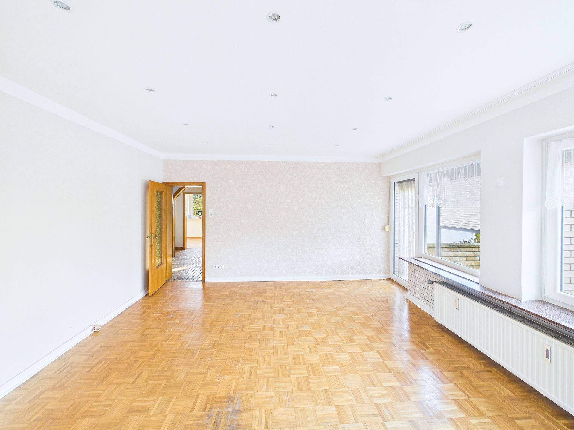 Heller, leerer Raum mit Parkettboden, großen Fenstern und einer offenen Holztür.