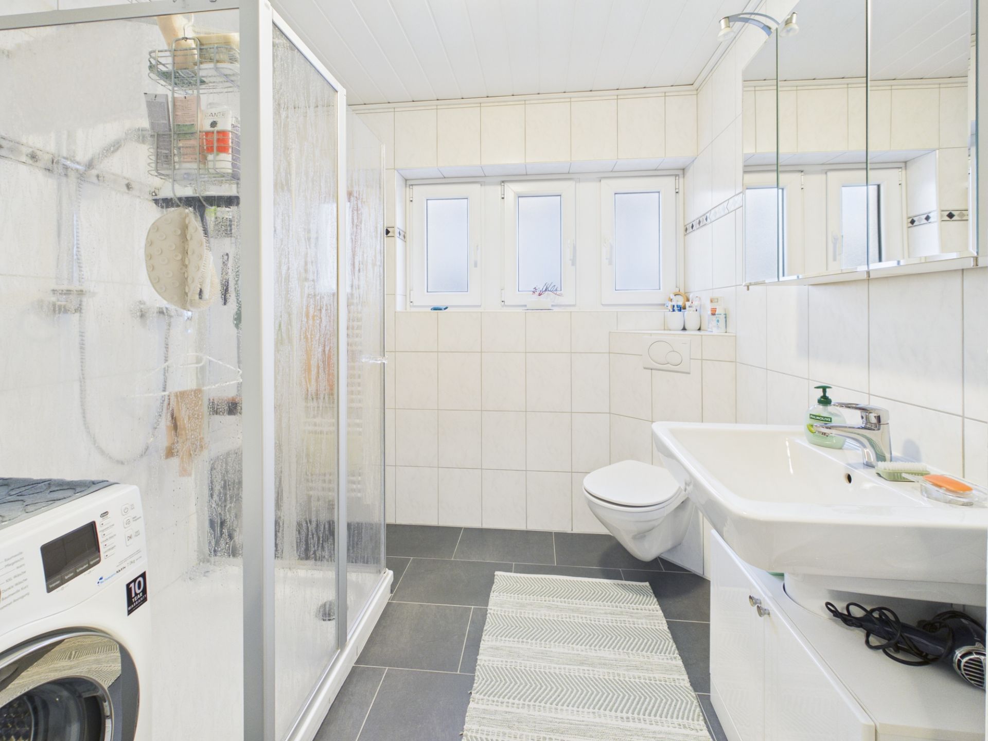 Helles Bad mit Dusche, Waschmaschine, Waschbecken, Toilette und drei Fenstern.
