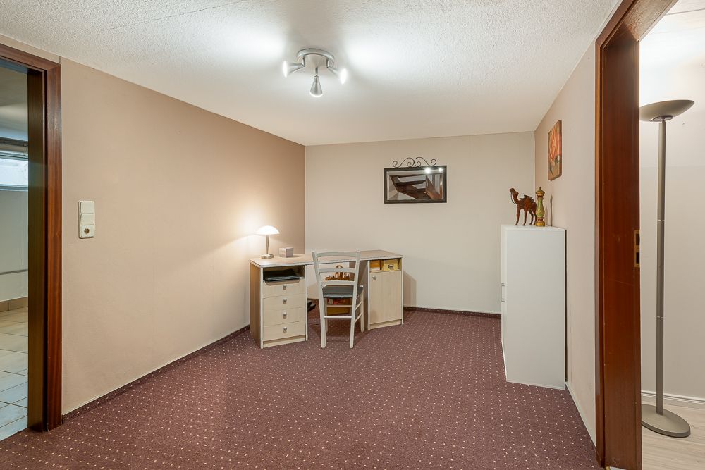 Kleines Zimmer mit Teppichboden, Schreibtisch, Stuhl, Lampe und Regal mit Dekoration.