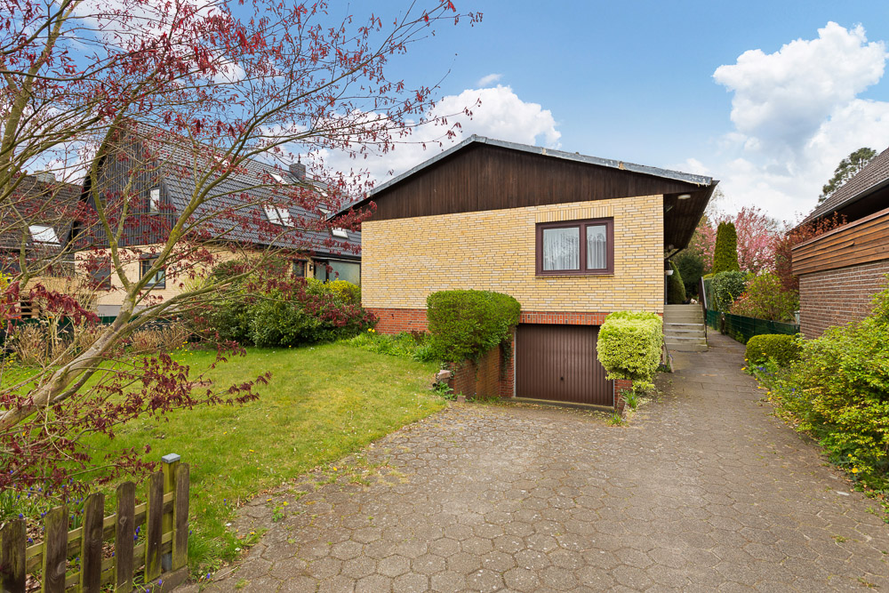 Einfamilienhaus mit gelber Klinkerfassade, Garage und Vorgarten mit Bäumen und Sträuchern.