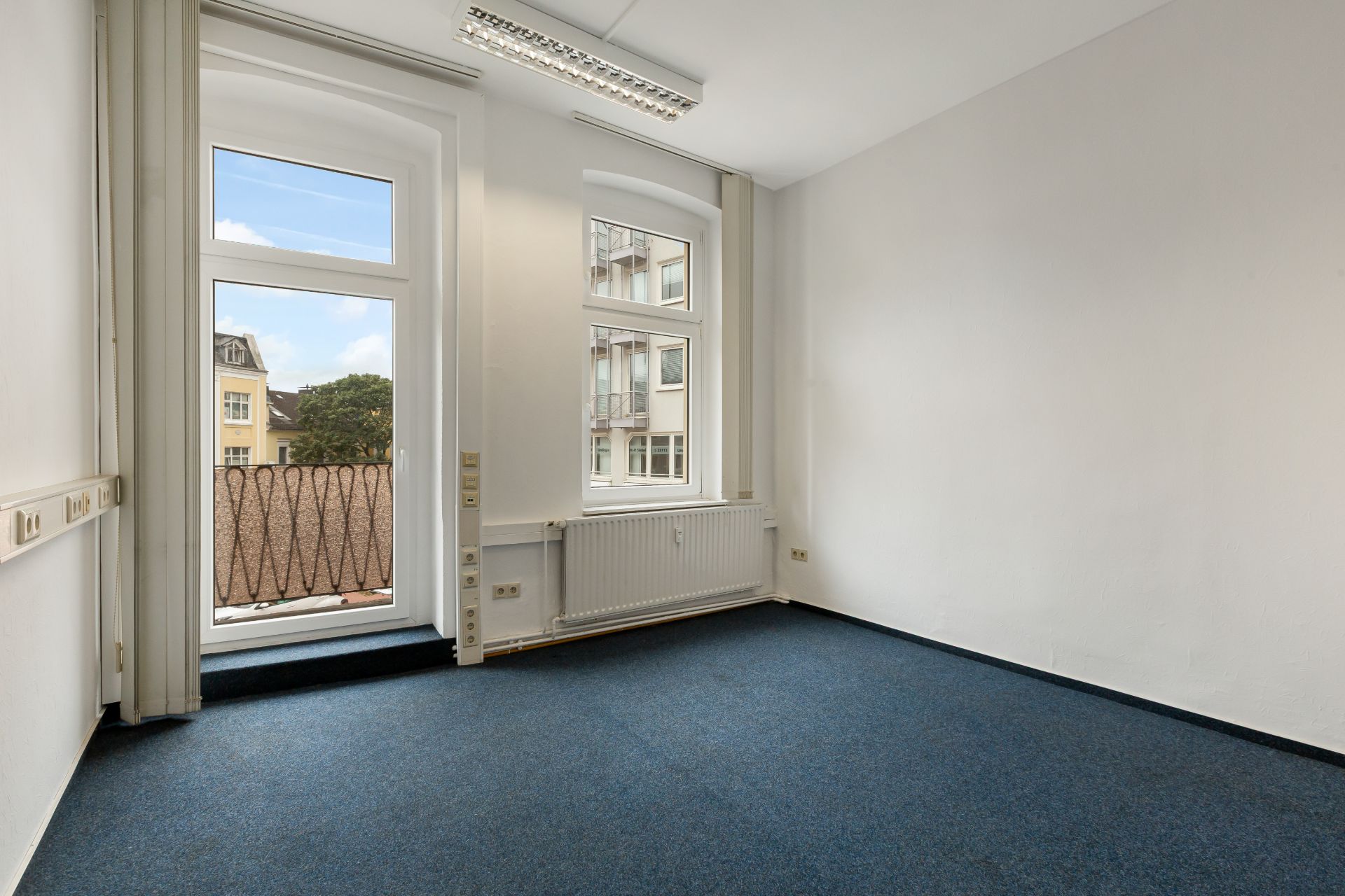 Heller Raum mit blauem Teppichboden, großen Fenstern und Heizkörper unter den Fenstern.