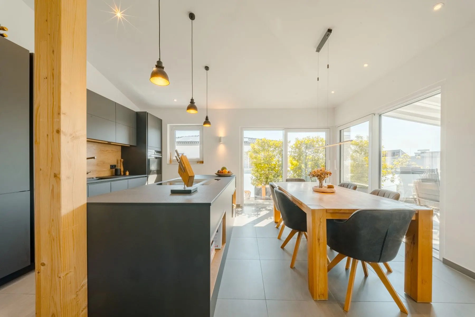 Moderne Küche mit Insel, Esstisch aus Holz und großen Fenstern mit Blick nach draußen.
