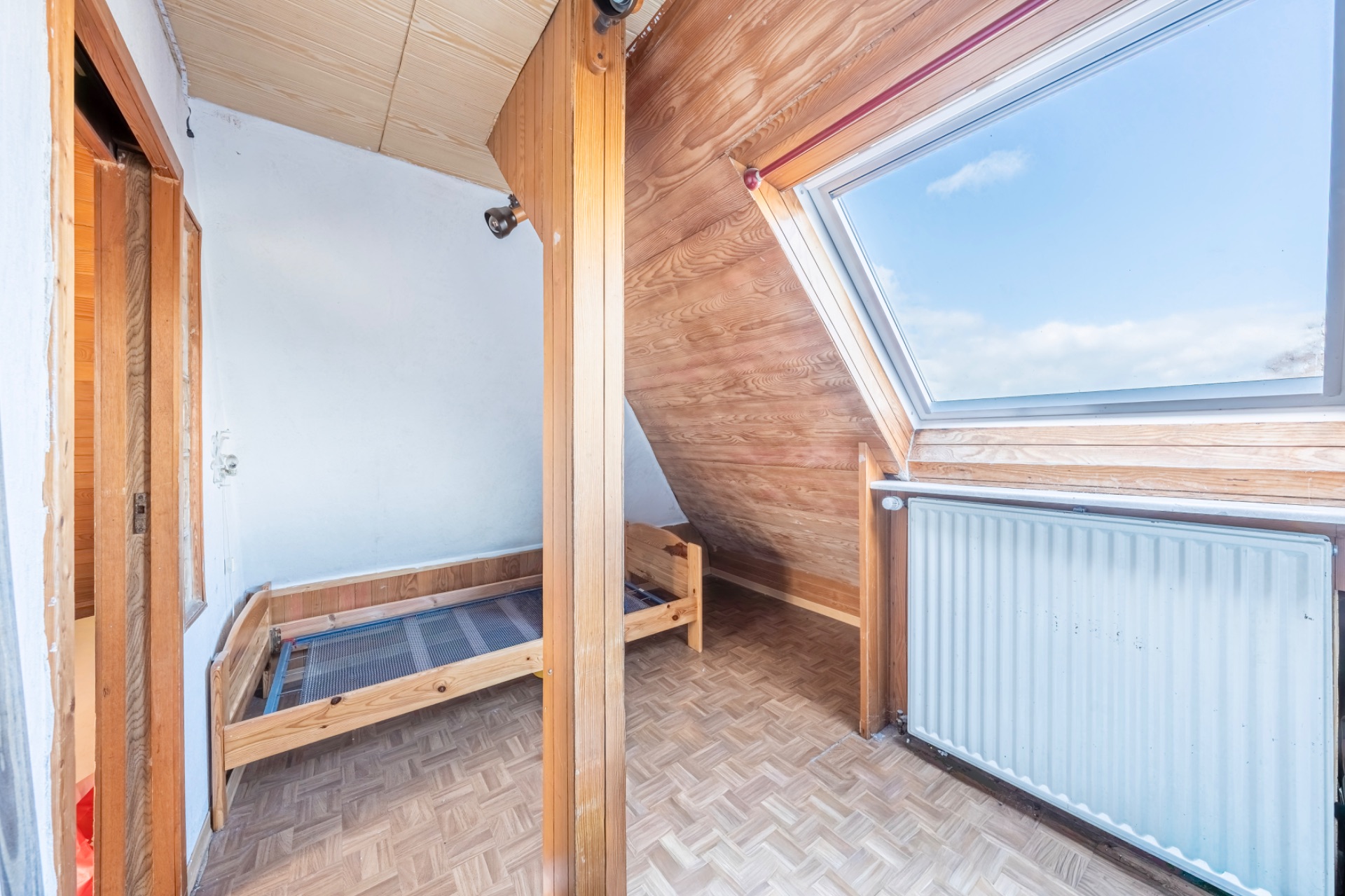 Kleines Zimmer mit Dachschräge, Bettgestell und großem Fenster mit Heizung darunter.