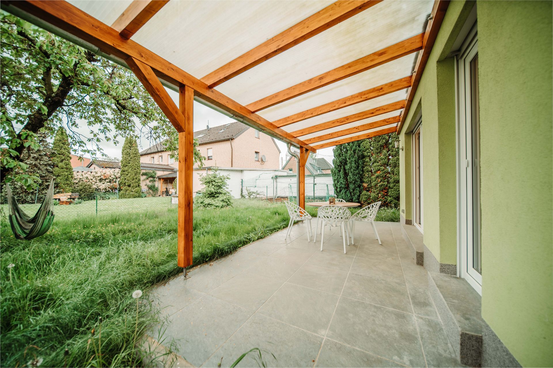 Überdachte Terrasse mit Tisch und Stühlen, daneben ein Garten mit Baum und Hängematte.