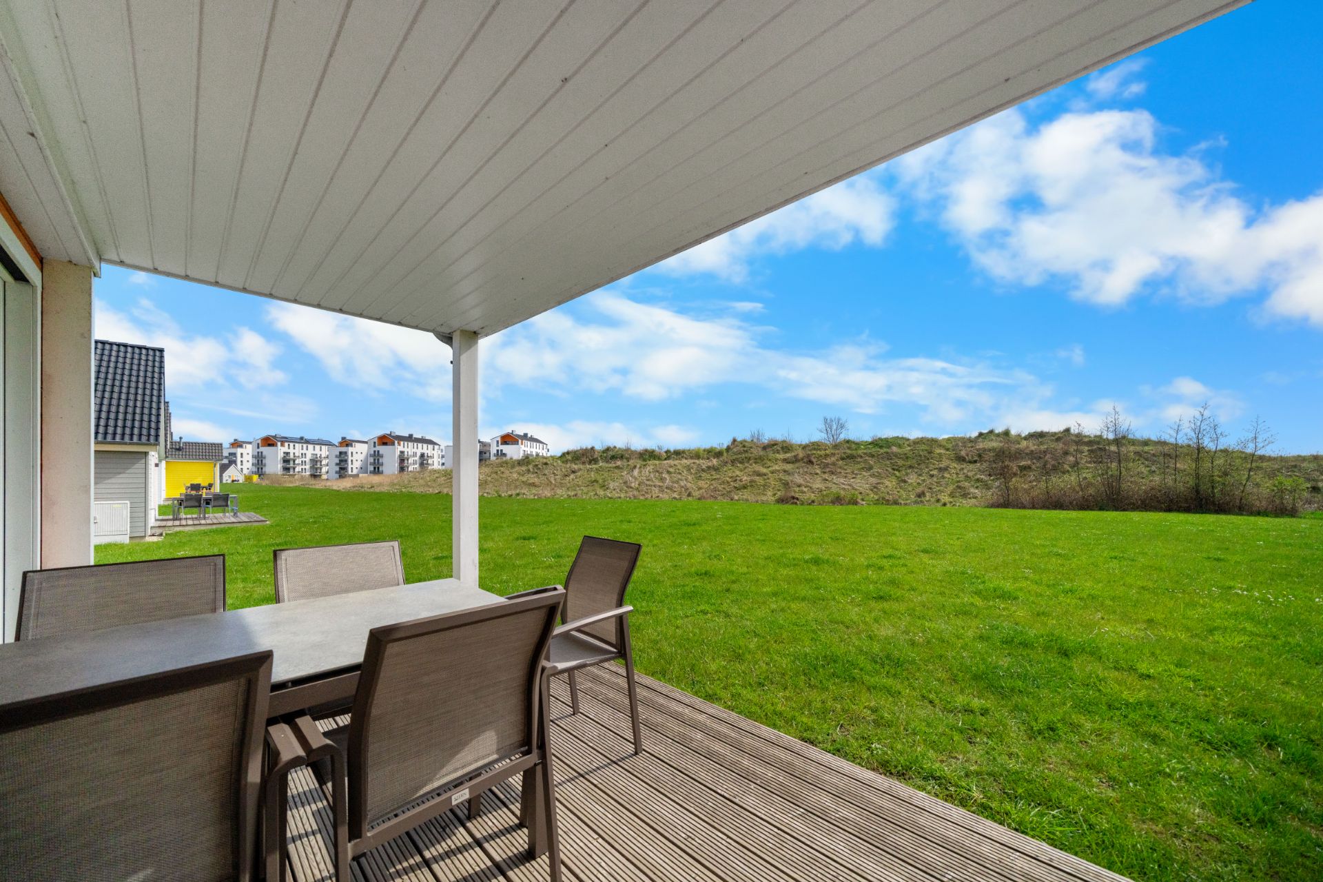 Überdachte Terrasse mit Esstisch und sechs Stühlen, Blick auf große Wiese und blauen Himmel.