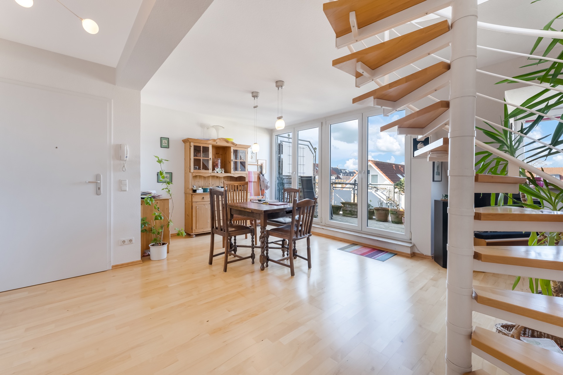 Helles Wohnzimmer mit Esstisch, Holzstühlen, Vitrine und Wendeltreppe vor großen Fenstern.