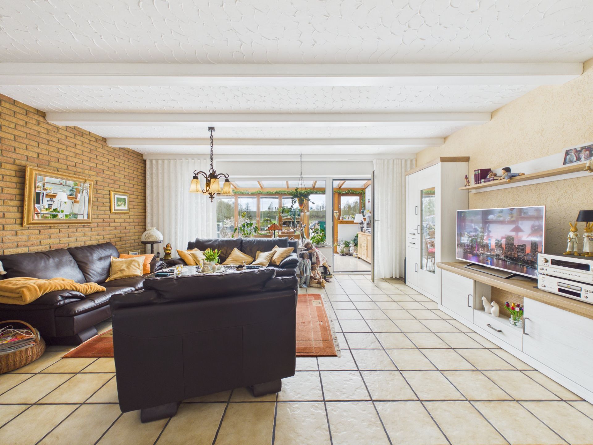 Helles Wohnzimmer mit Ledersofas, Fernseher, gemauerter Wand und Zugang zum Wintergarten.