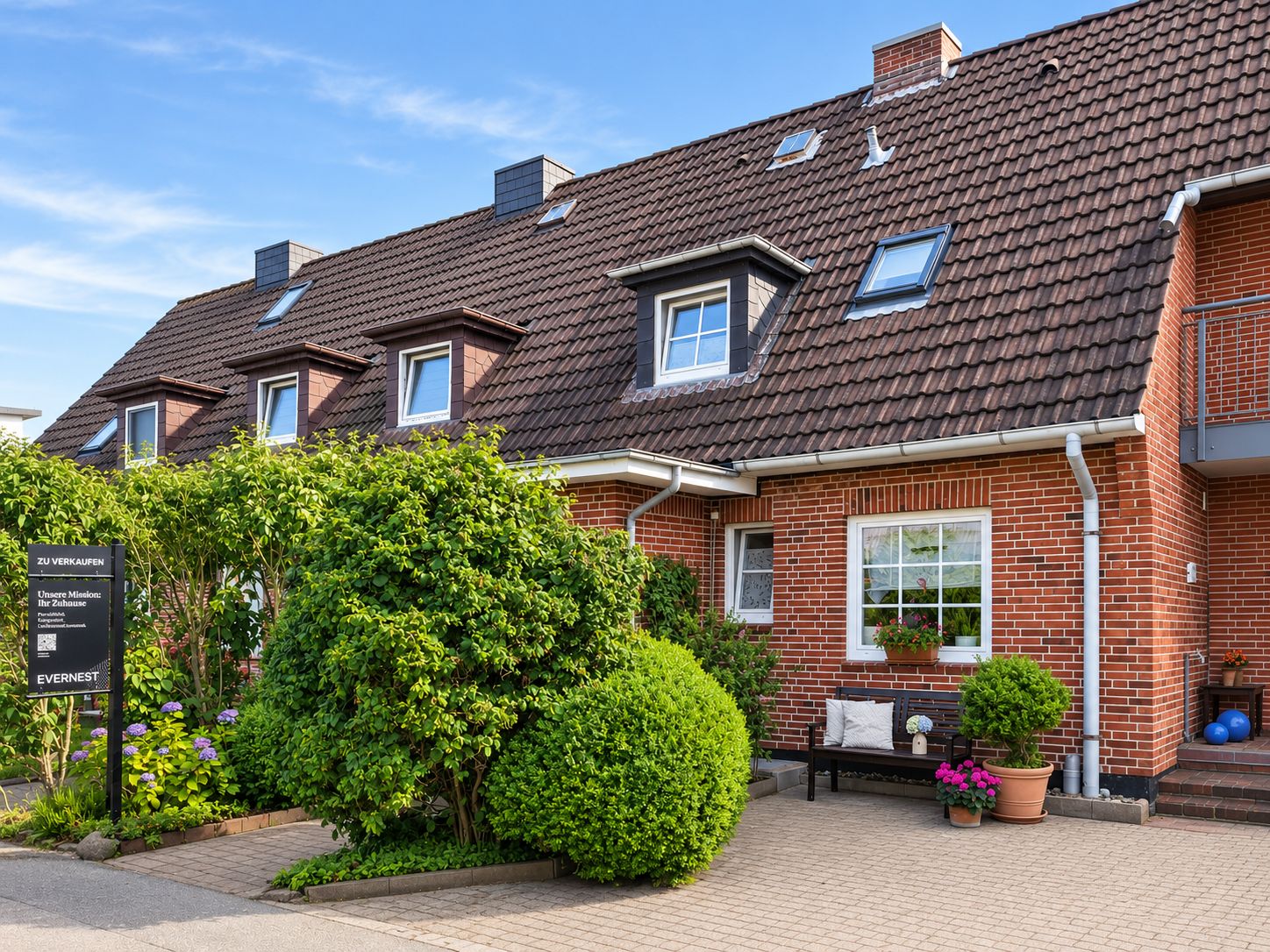 Reihenhaus mit rotem Klinker, braunem Dach und gepflegtem Vorgarten mit Sträuchern und Bank.