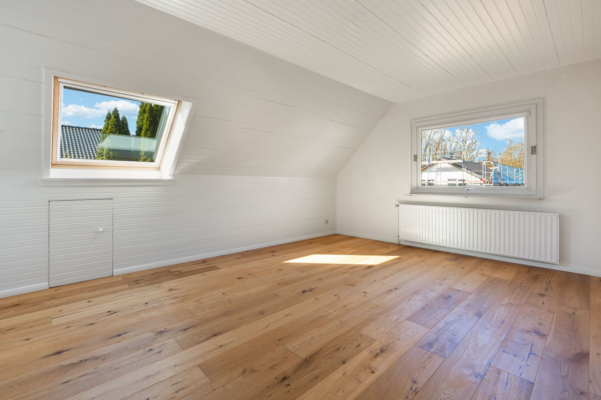 Helles Dachgeschosszimmer mit Holzboden, zwei Fenstern und weiß gestrichenen Wänden.