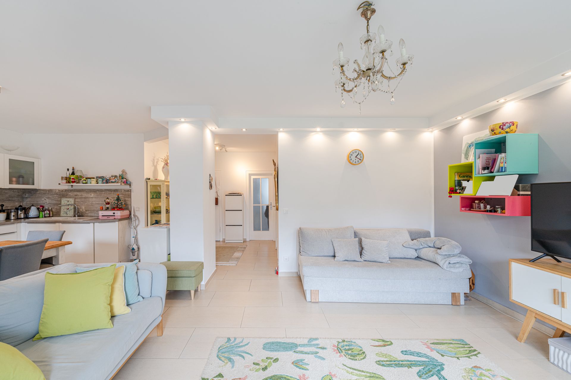 Helles Wohnzimmer mit Sofa, Teppich, Küche im Hintergrund und bunten Regalen an der Wand.