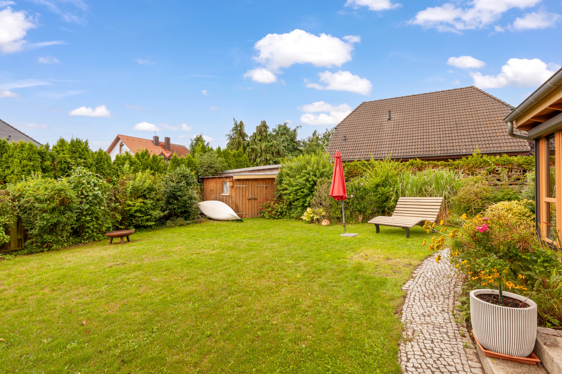 Grüner Garten mit Rasen, Bank, rotem Sonnenschirm, Gartenhaus und Pflanzen.