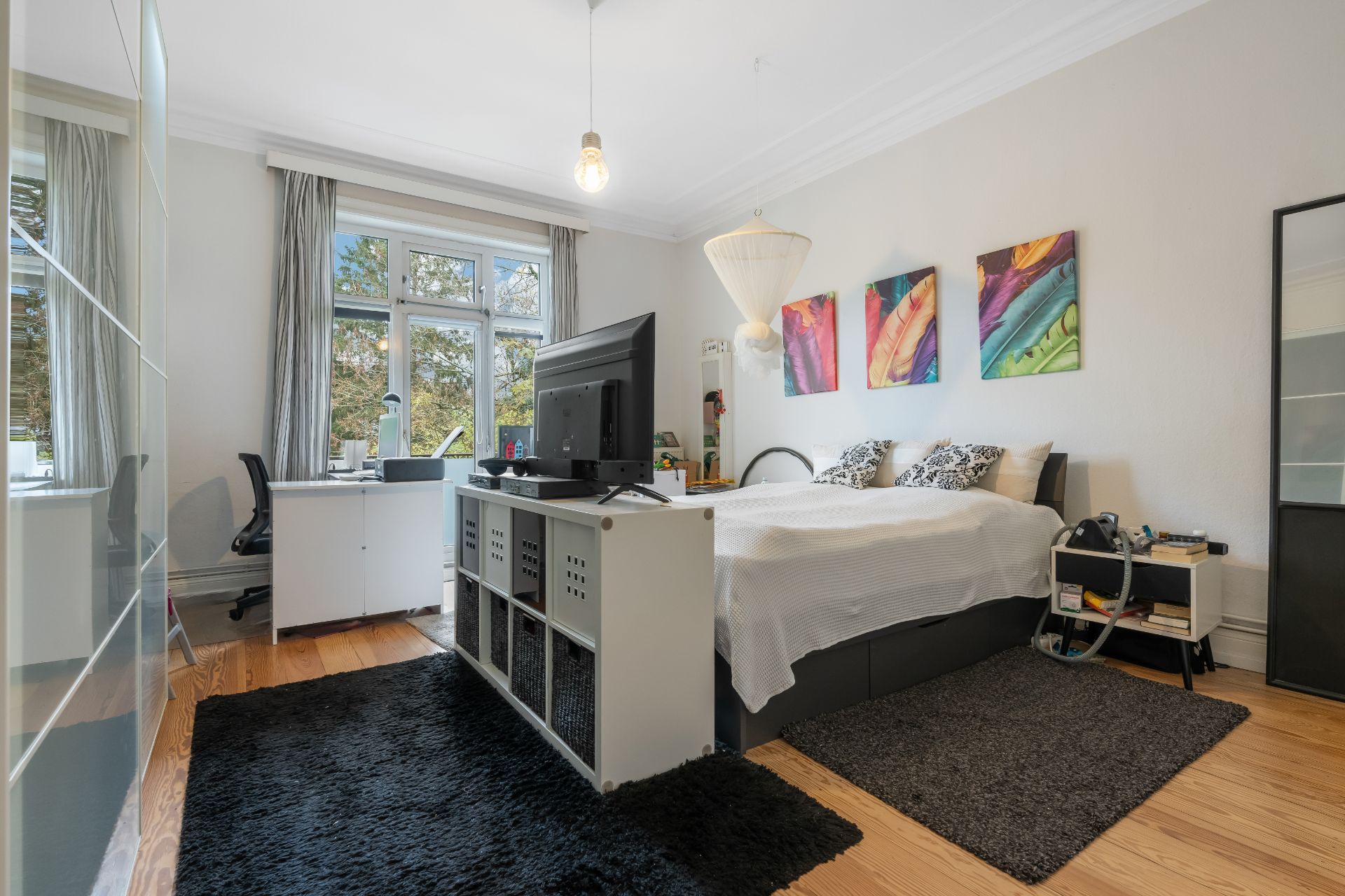 Helles Schlafzimmer mit großem Bett, Schreibtisch, Fernseher und bunten Bildern an der Wand.