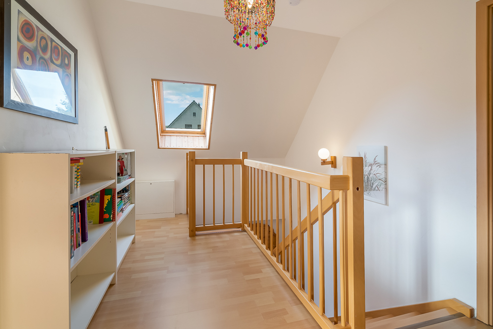 Heller Flur mit Bücherregal, Holzgeländer, buntem Kronleuchter und Dachfenster.