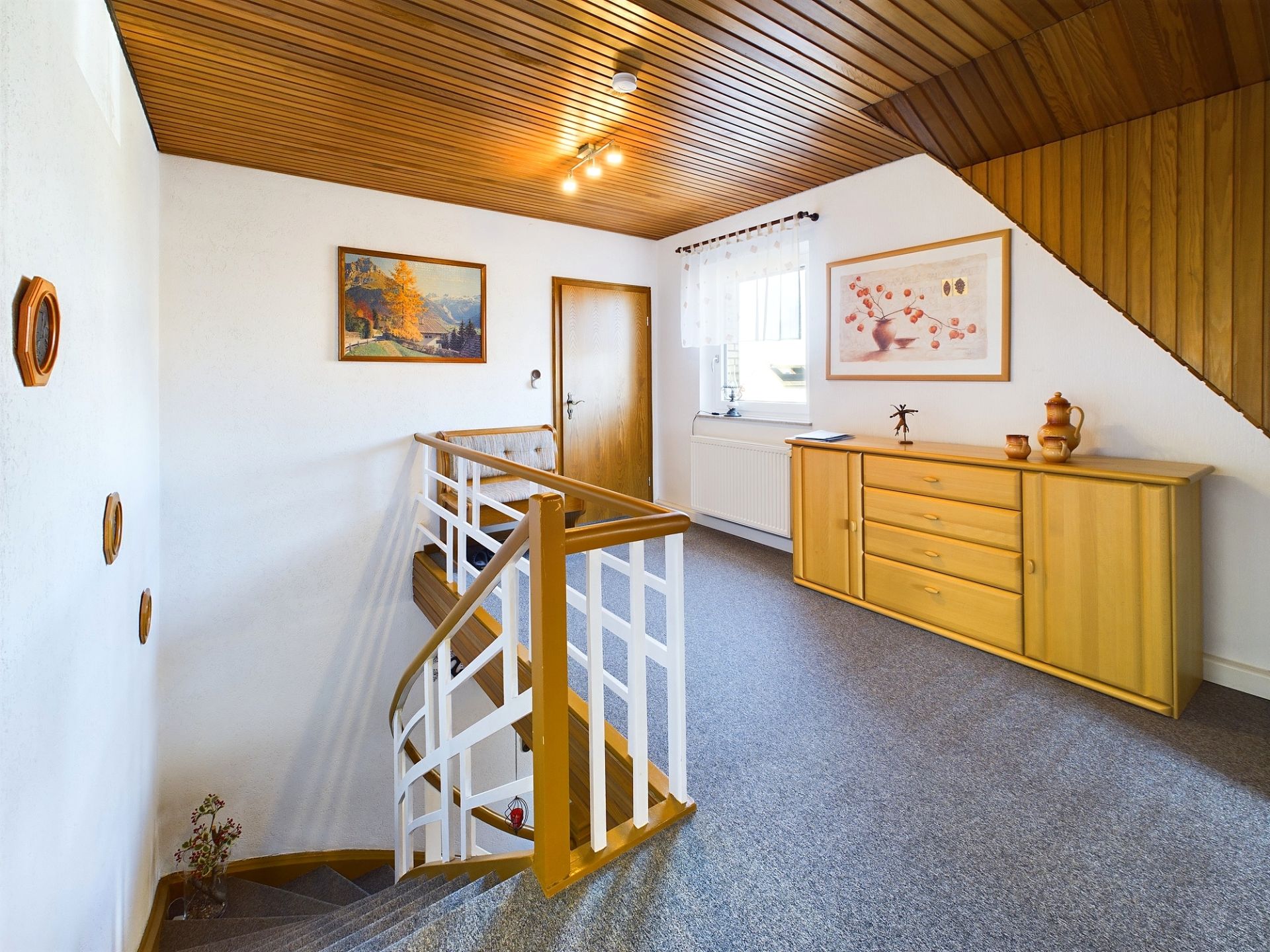 Heller Flur mit Holzdecke, Treppe, Kommode und Bildern an der Wand.