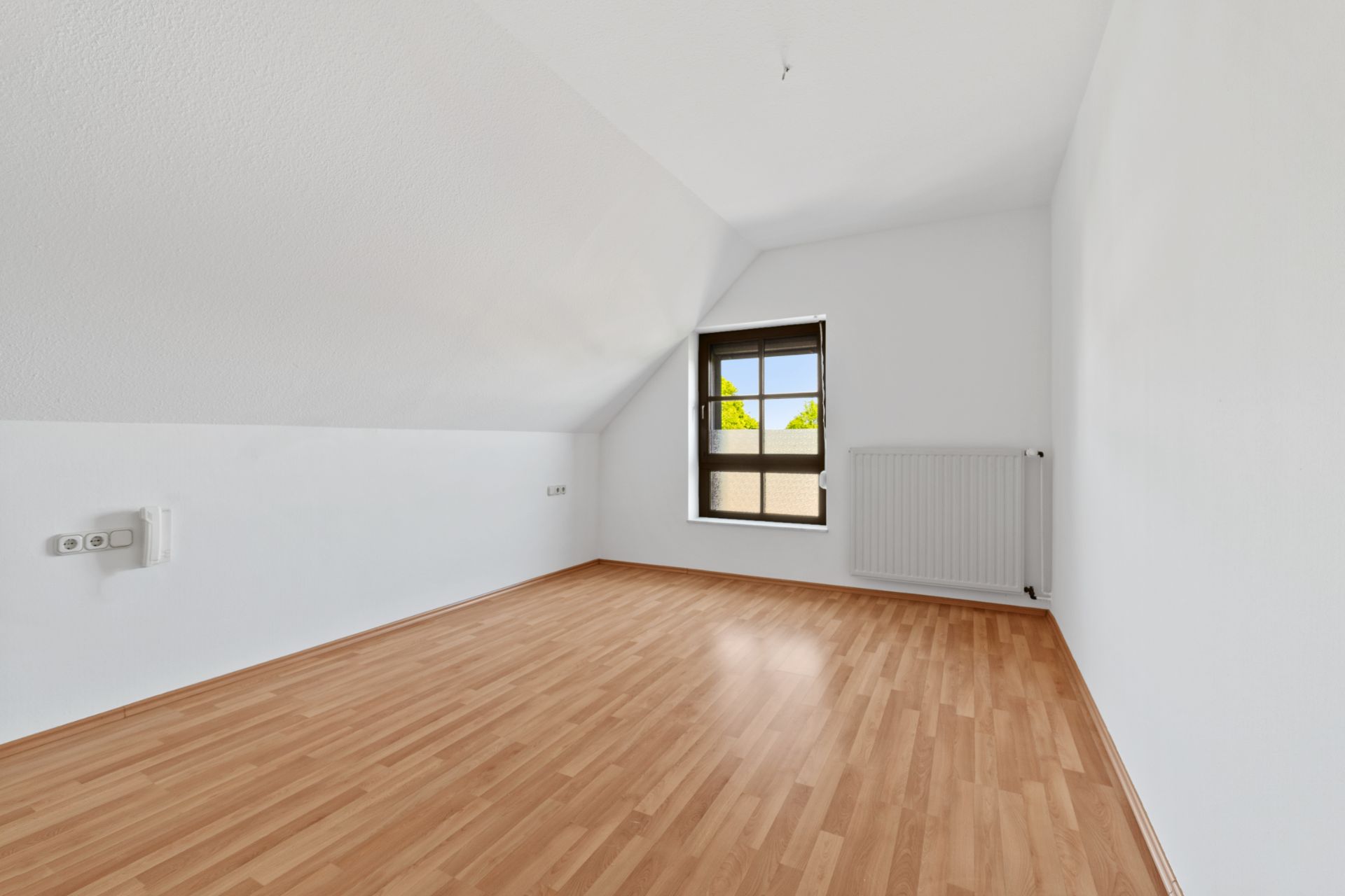 Leerer Raum mit Holzfußboden, weiß gestrichenen Wänden und einem Fenster mit Heizung darunter.