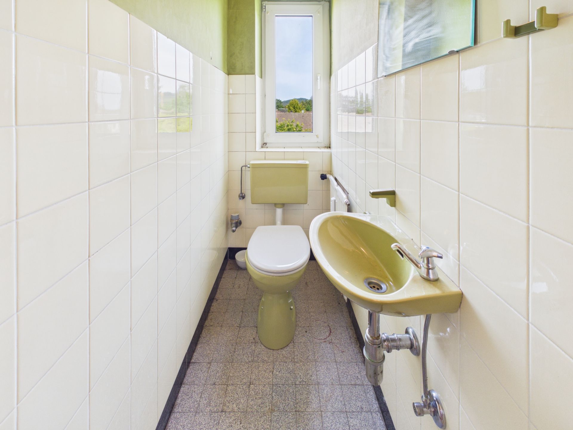 Schmales Badezimmer mit Toilette, kleinem Waschbecken und Fenster am Ende.