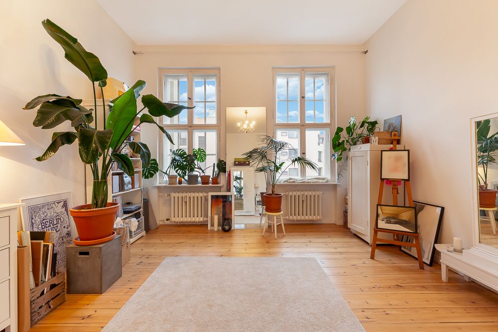 Helles Wohnzimmer mit Holzboden, großen Pflanzen, Teppich und zwei Fenstern.