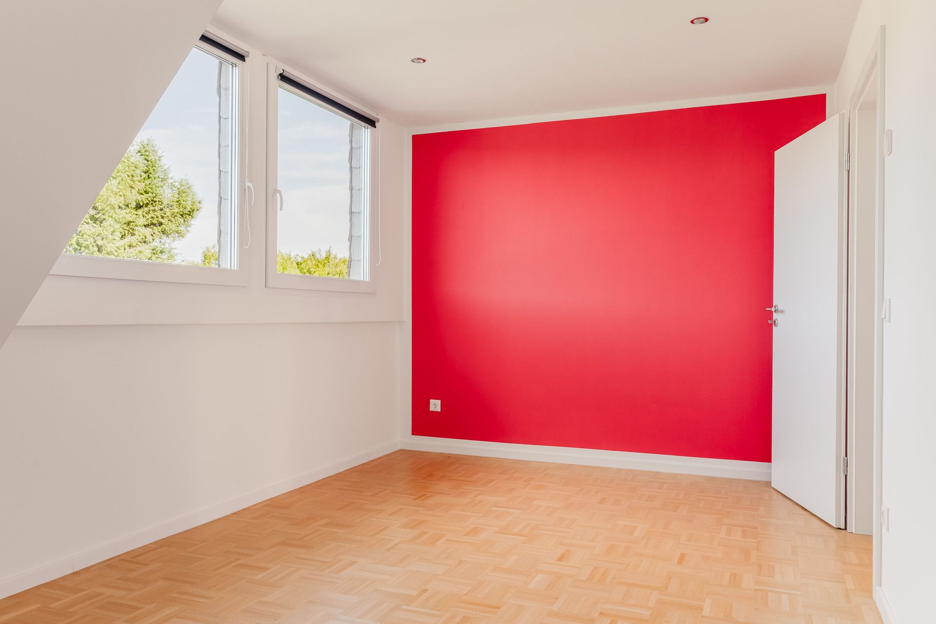 Leeres Zimmer mit roter Wand, Parkettboden und zwei Fenstern auf der linken Seite.