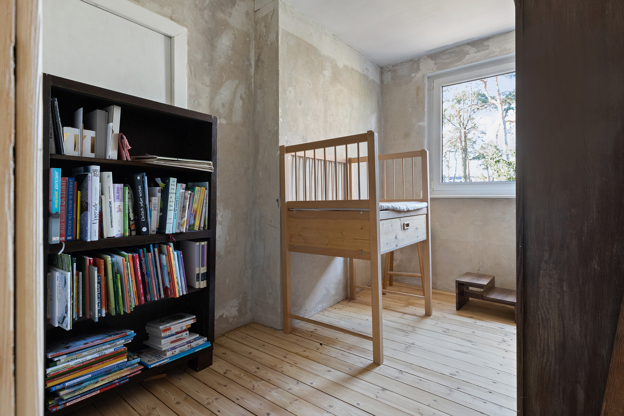 Kleines Zimmer mit Holzfußboden, Bücherregal und Kinderbett am Fenster.