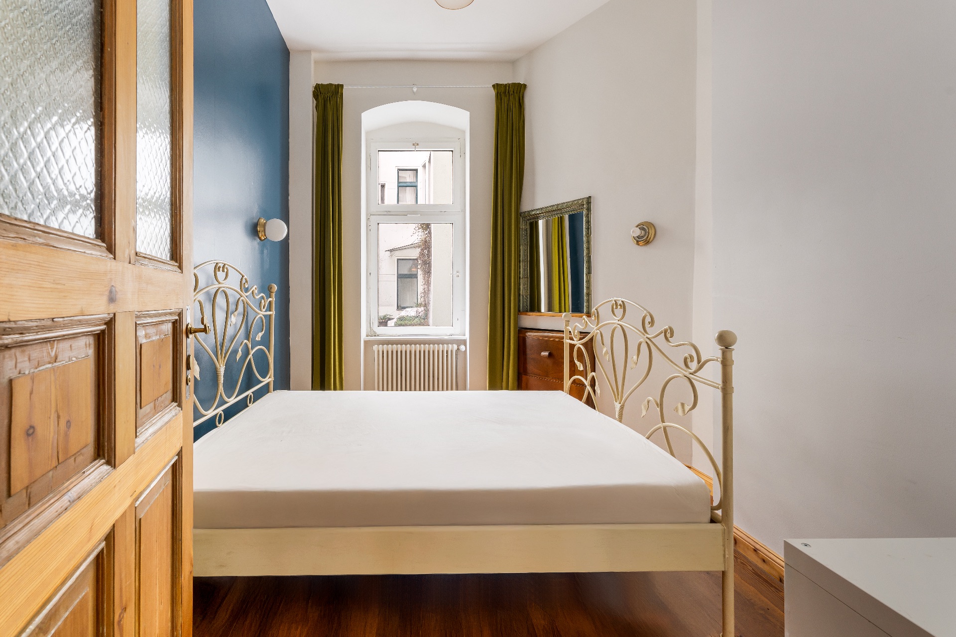 Helles Schlafzimmer mit Metallbett, Holzboden, grünem Vorhang und Fenster zur Straße.