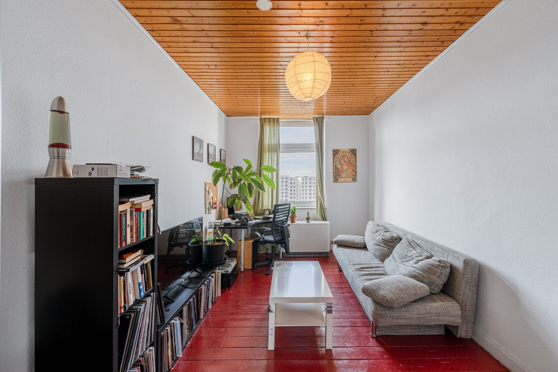 Wohnzimmer mit rotem Holzboden, grauem Sofa, Bücherregal, Schreibtisch und Fenster mit grünen Vorhängen.