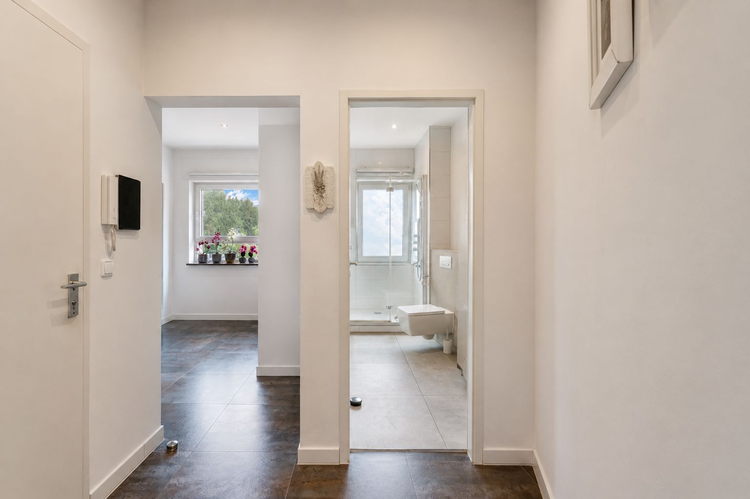 Flur mit Blick ins Badezimmer rechts und in einen Raum mit Fenster und Blumen links.
