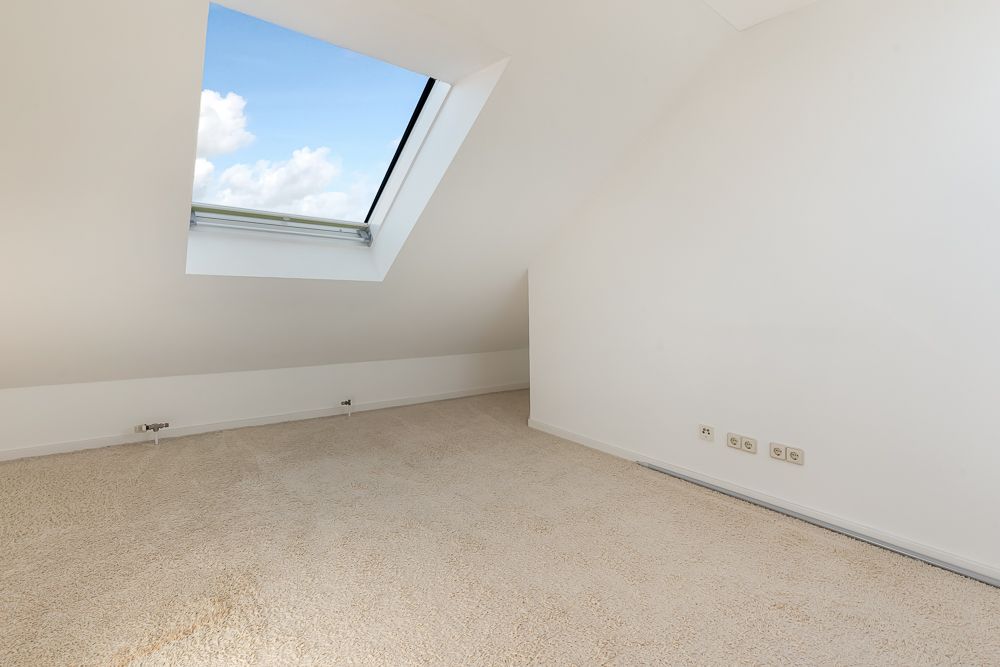 Leerer Raum mit Teppichboden und Dachfenster, weiße Wände, Blick auf blauen Himmel.