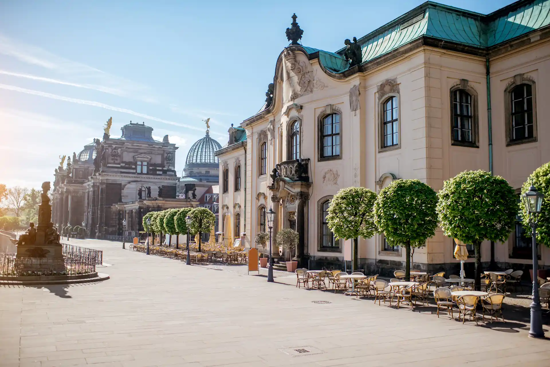 Historisches Gebäude mit Außenterrasse, Tischen, Stühlen und Bäumen entlang einer breiten gepflasterten Straße.