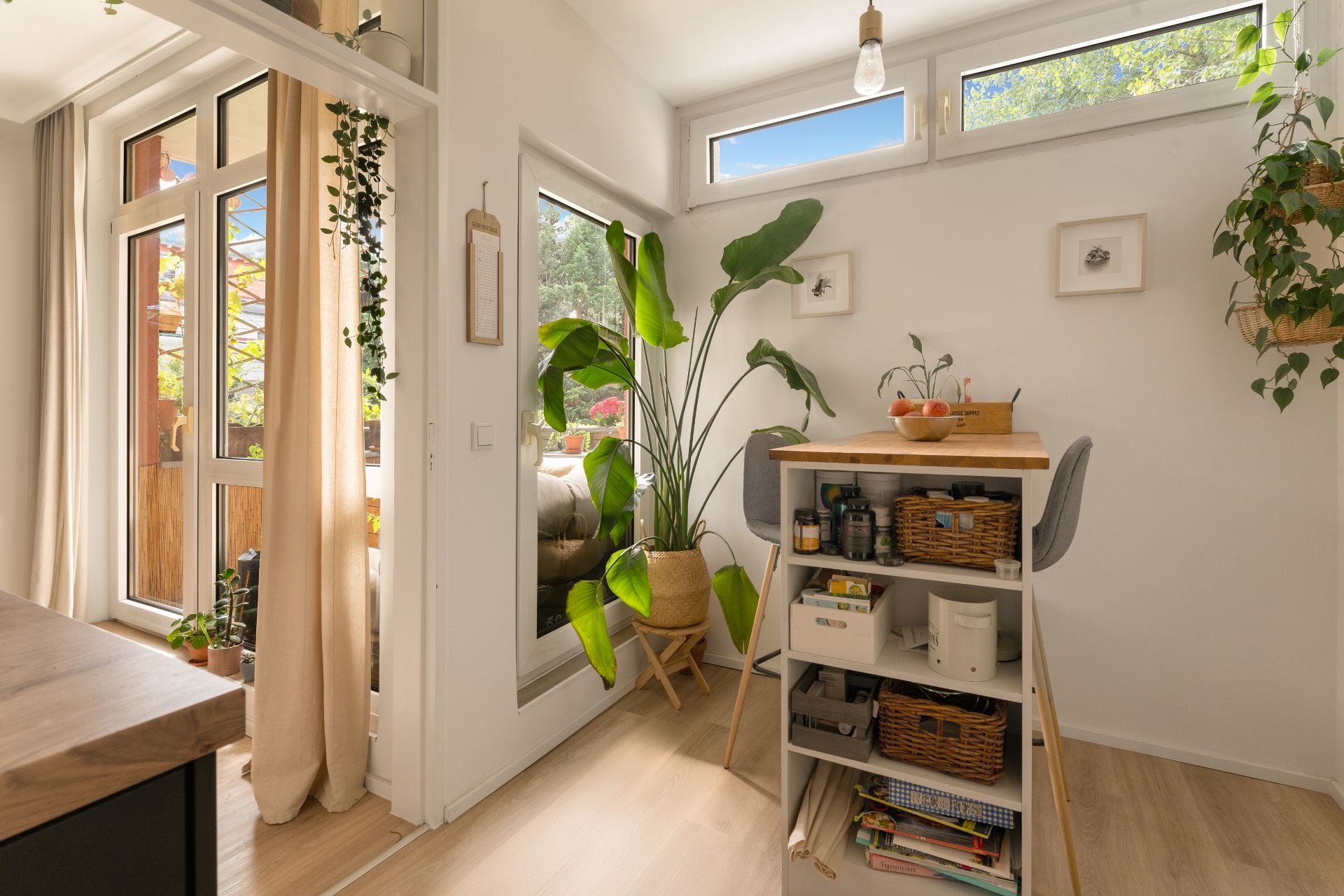 Helles Zimmer mit Regal, Pflanzen, Tisch und Zugang zu einem Balkon mit Vorhängen.