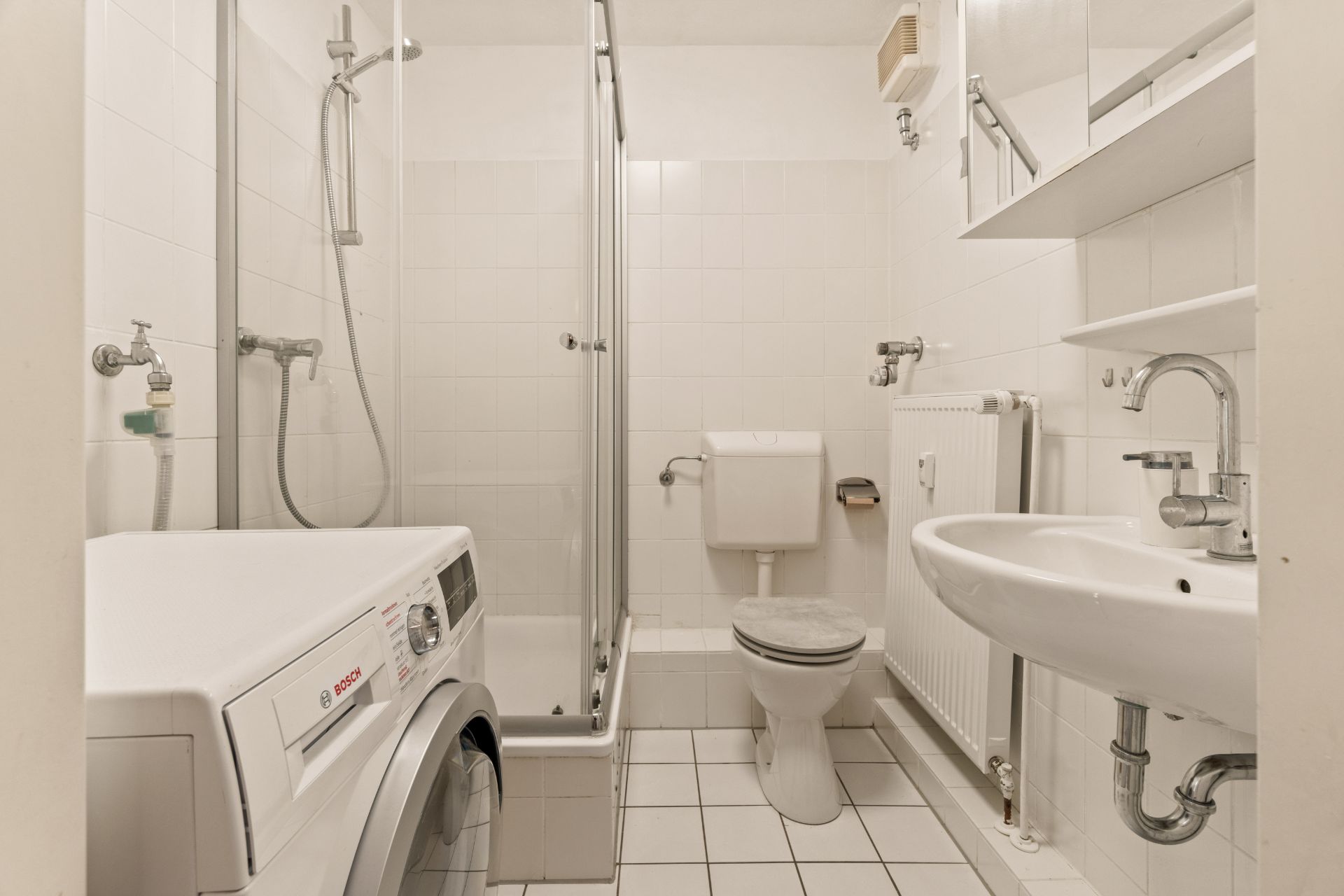 Helles Badezimmer mit Dusche, Waschmaschine, Toilette, Waschbecken und weißen Fliesen.