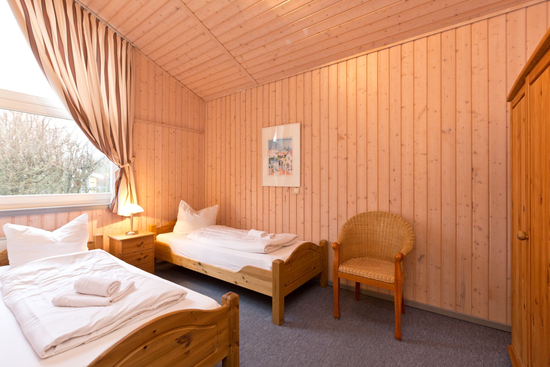 Schlafzimmer mit zwei Einzelbetten, Holzwänden, Stuhl, Schrank und Fenster mit Vorhang.