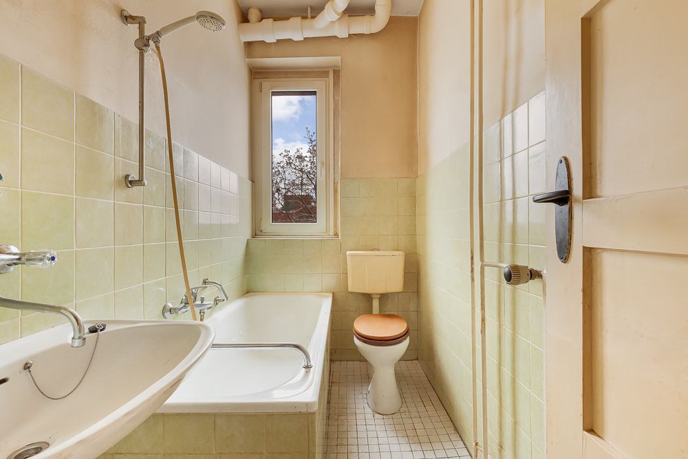 Kleines Badezimmer mit Badewanne, Waschbecken, Toilette und Fenster, grüne Fliesen an den Wänden.