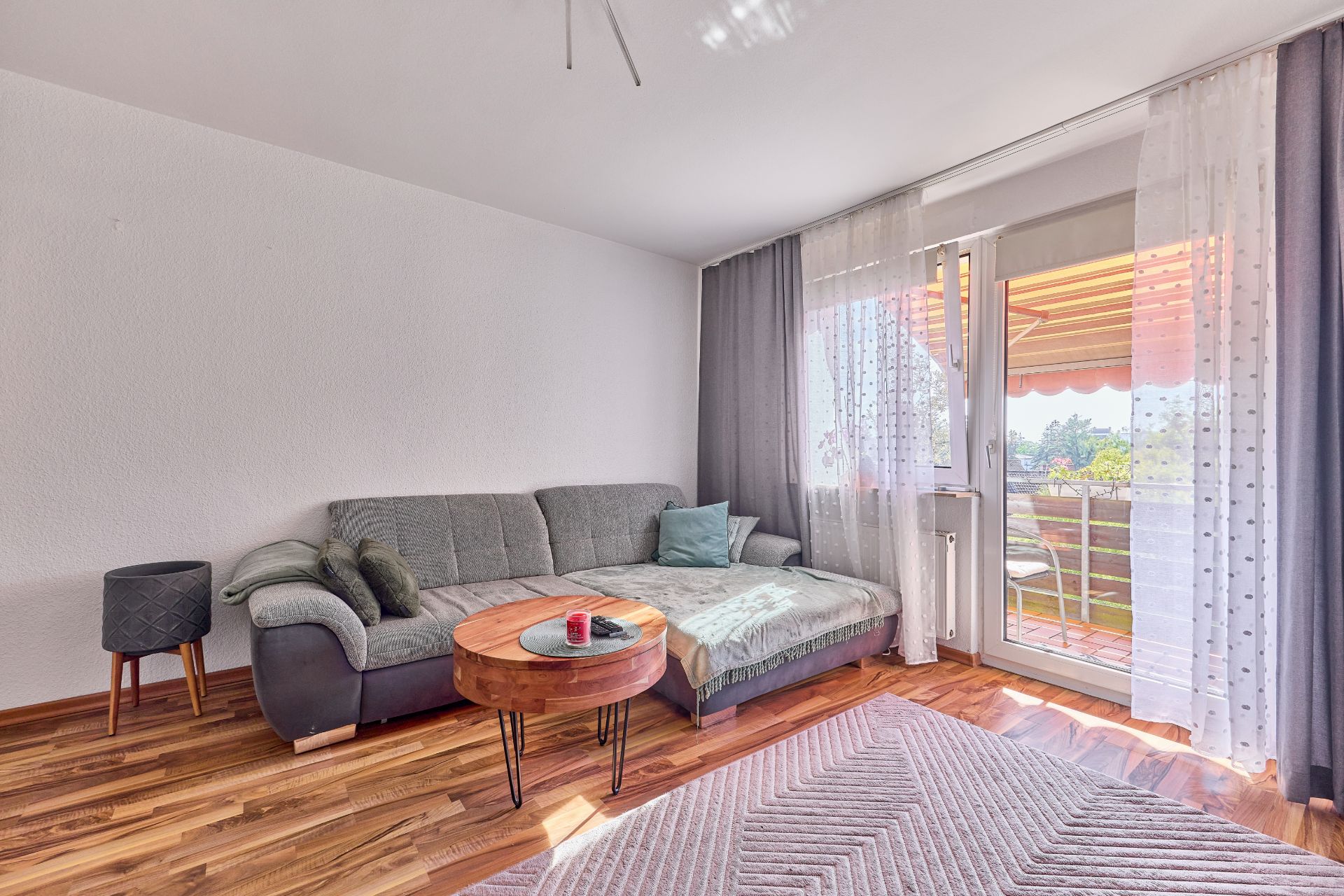 Wohnzimmer mit grauem Sofa, rundem Holztisch und Zugang zum Balkon, helle Vorhänge am Fenster.