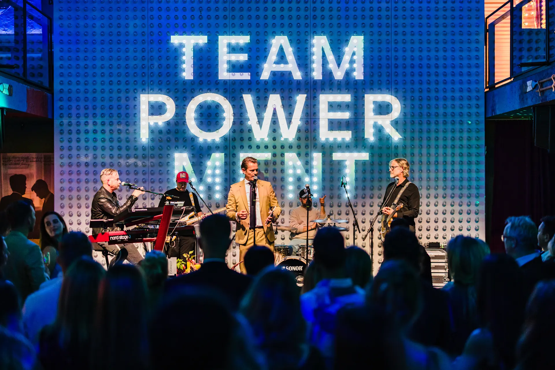 Eine Musikband spielt auf einer Bühne vor Publikum, Hintergrund mit großem Schriftzug TEAM POWER MENT.