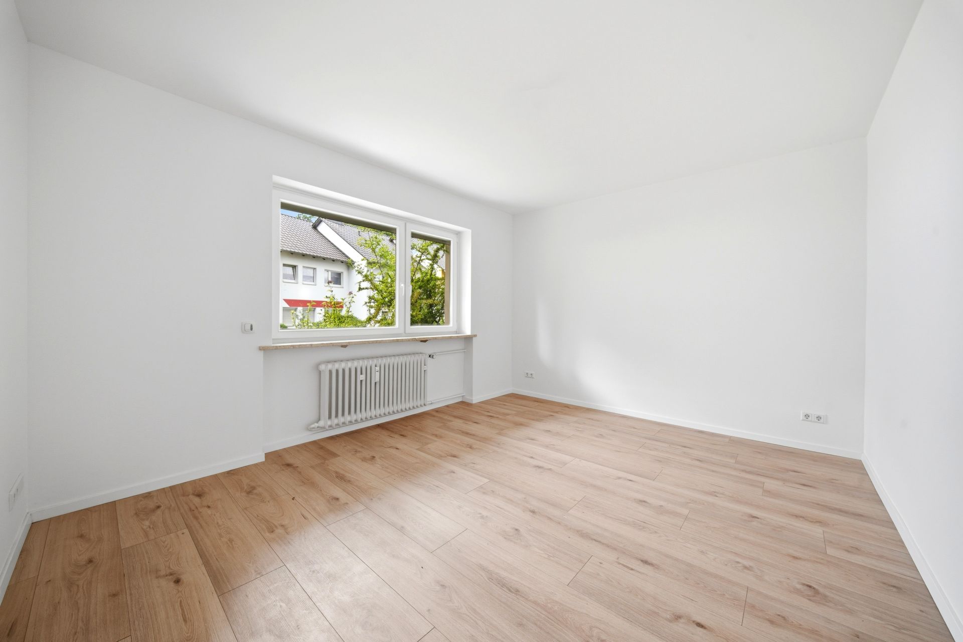 Leerer, heller Raum mit Holzboden, weiß gestrichenen Wänden und großem Fenster mit Blick nach draußen.