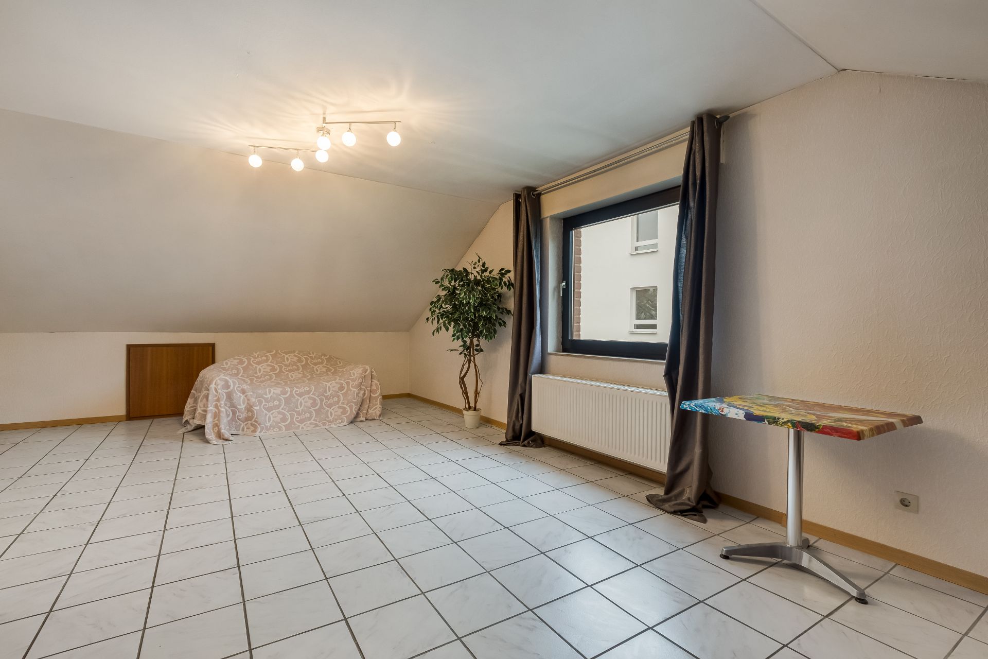 Helles Zimmer mit schräger Decke, Fliesenboden, Couch, Pflanze, Fenster und kleinem Tisch.