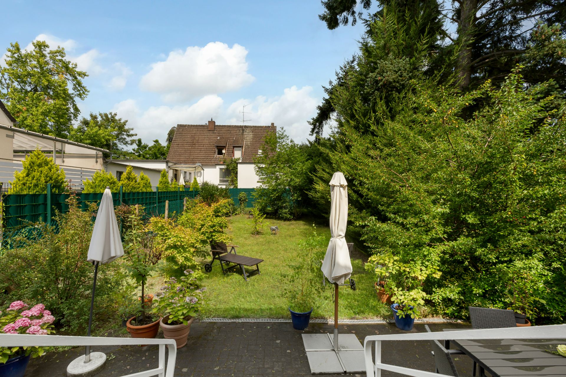Blick auf einen grünen Garten mit Terrasse, Pflanzen, Sonnenschirmen und Liegestuhl.