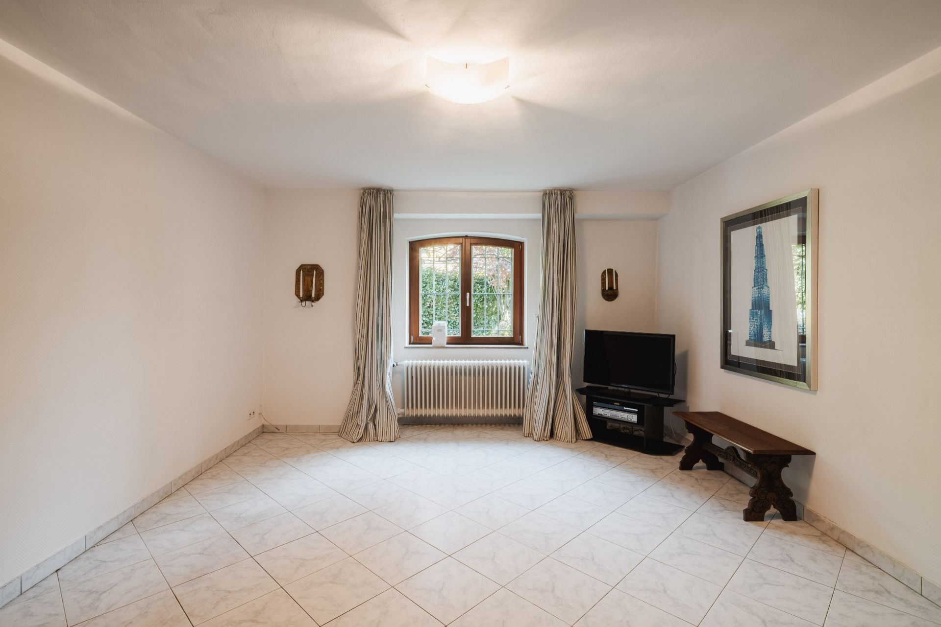 Helles Wohnzimmer mit Fliesenboden, Fenster, Fernseher, Bank und Wandbild.