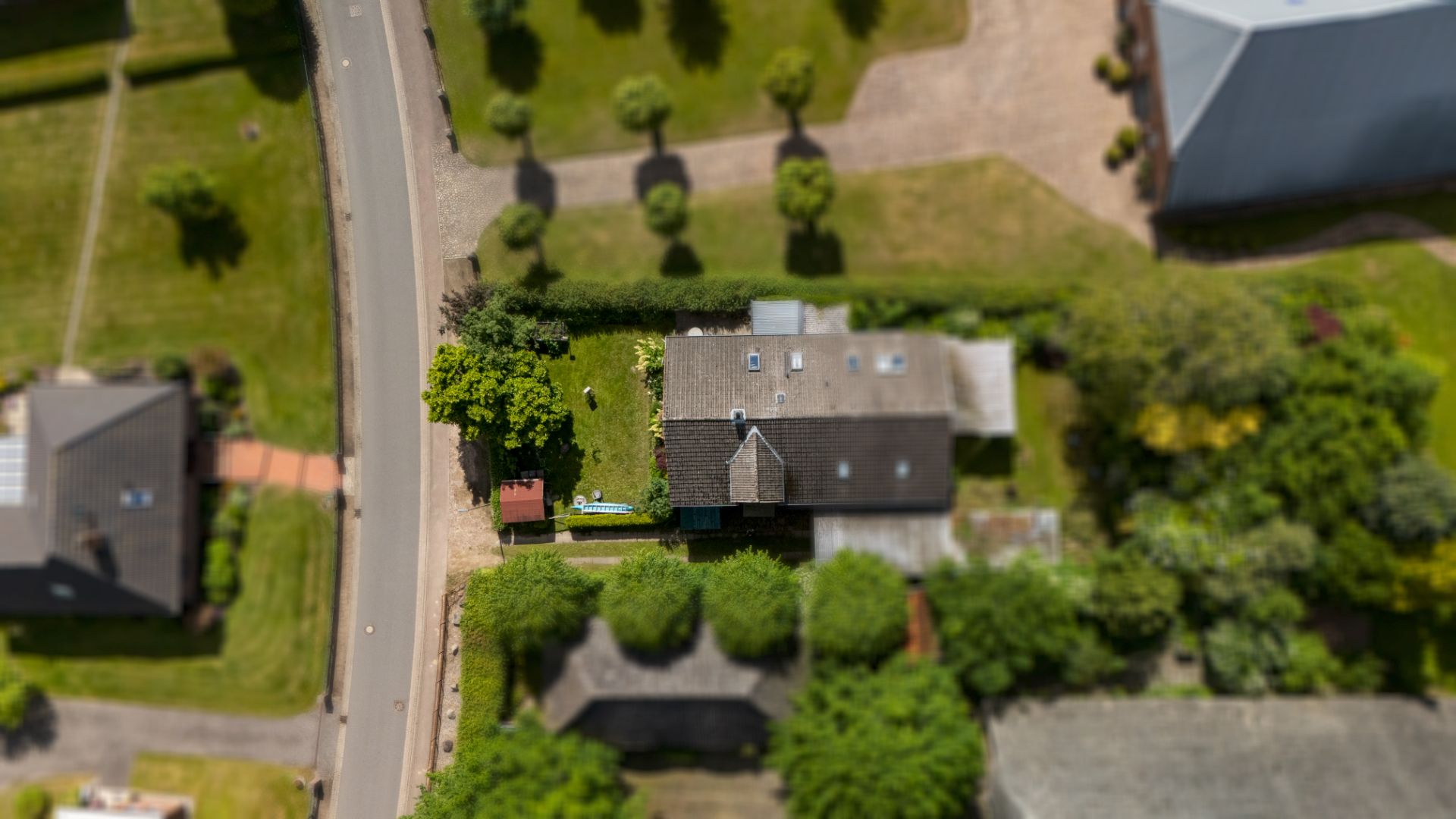 Luftaufnahme eines Wohnhauses mit Garten an einer Straße, umgeben von Bäumen und Nachbargebäuden.