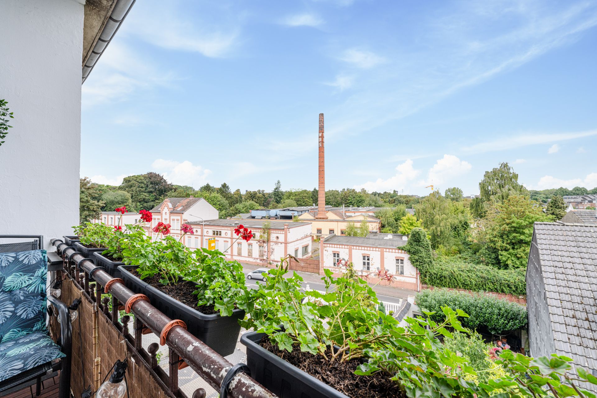 Balkon mit Blumenkästen, Blick auf alte Gebäude und einen Schornstein, umgeben von Bäumen.