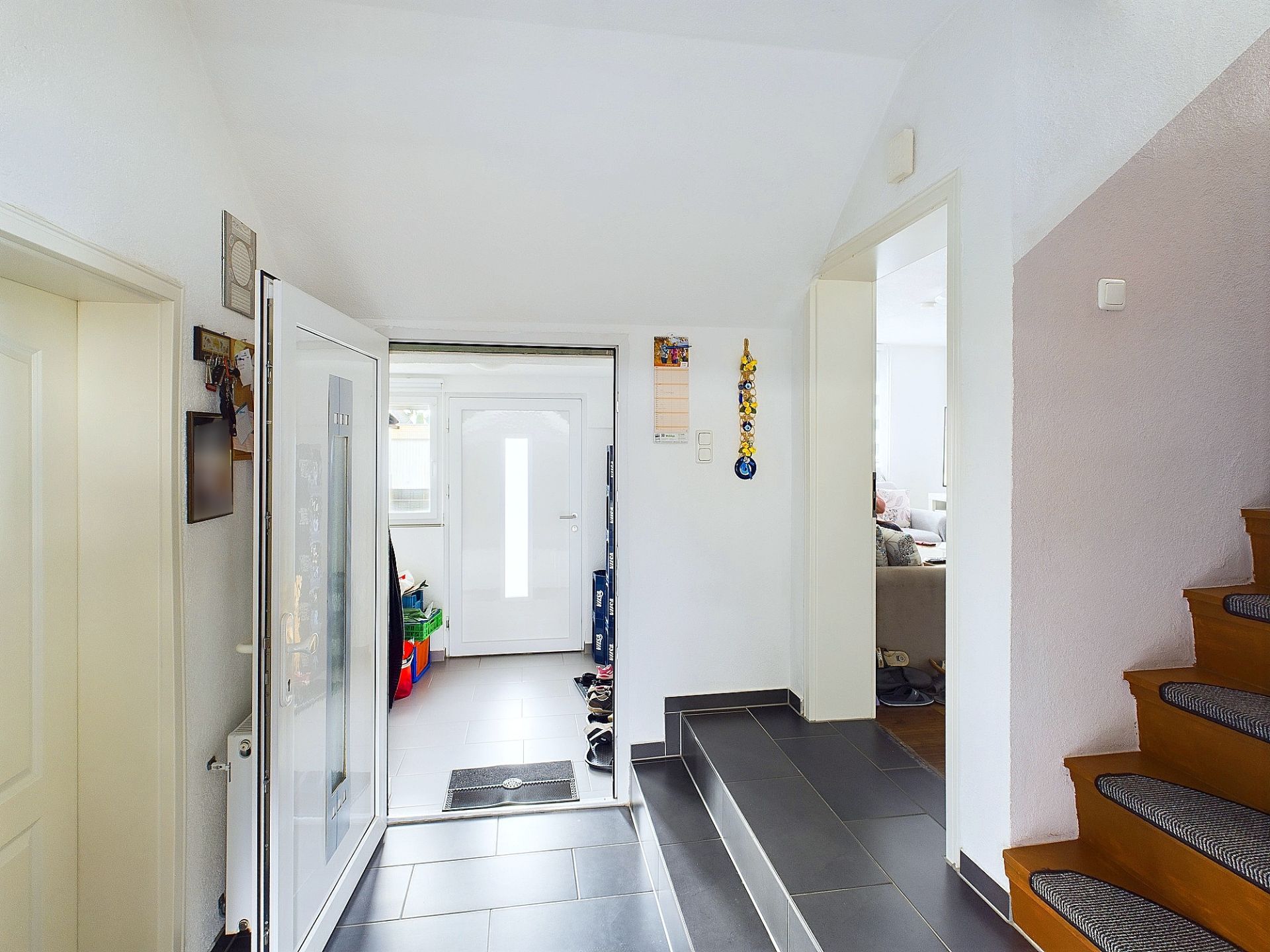 Heller Flur mit grauen Fliesen, Treppe, offenem Eingang und Blick ins Wohnzimmer.
