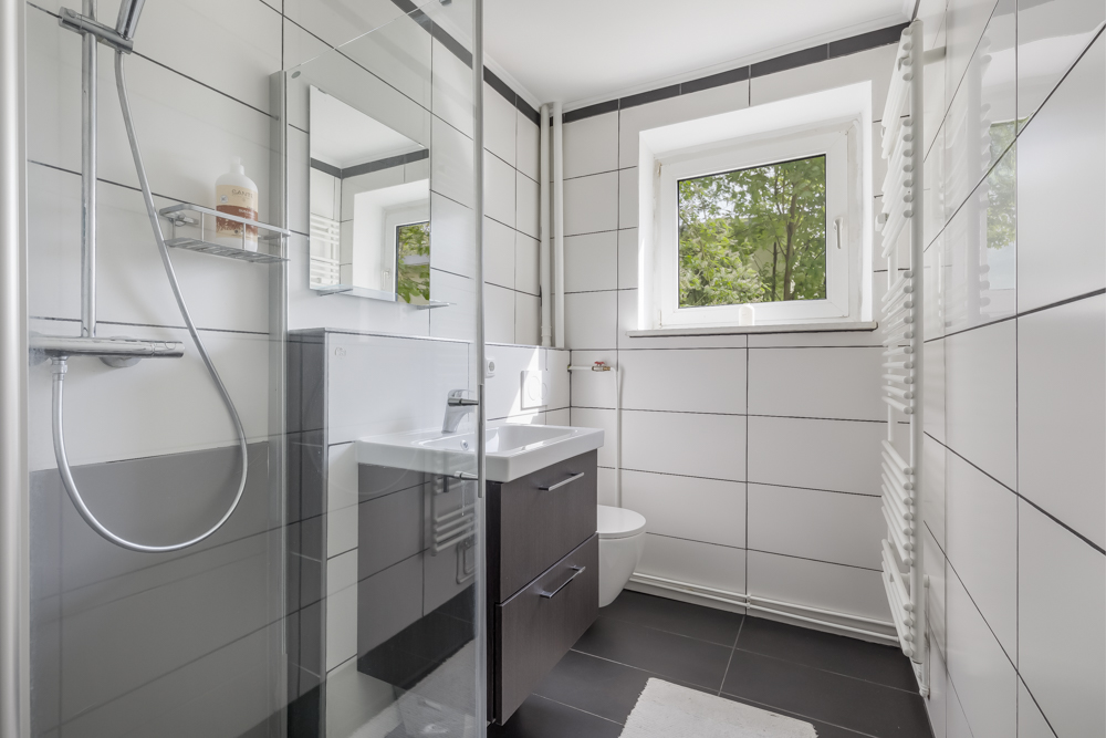 Modernes Bad mit Dusche, Waschbecken, Spiegel, Fenster und WC, weiße Fliesen.
