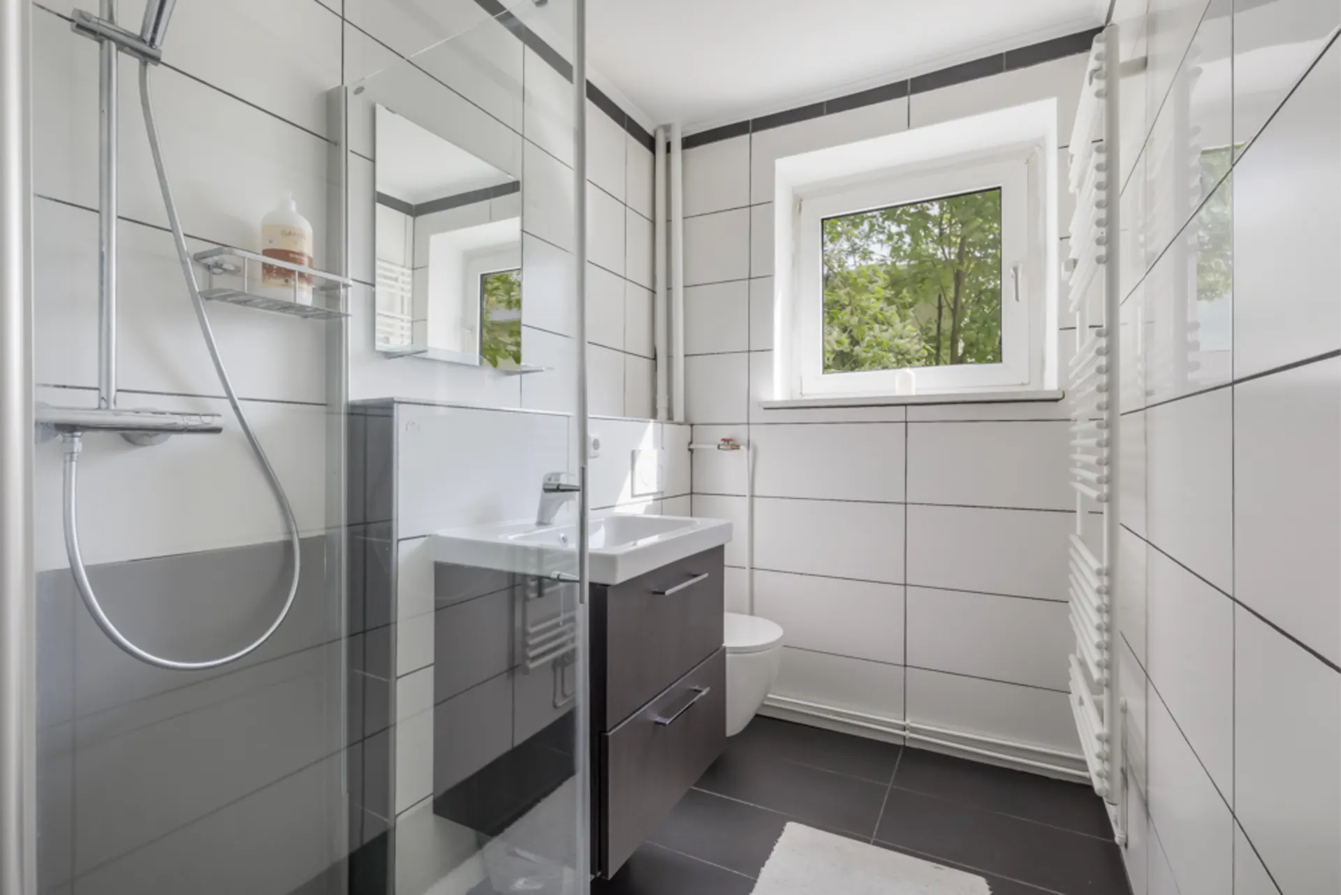 Modernes Bad mit Dusche, Waschbecken, Spiegel, Fenster und WC, weiße Fliesen.