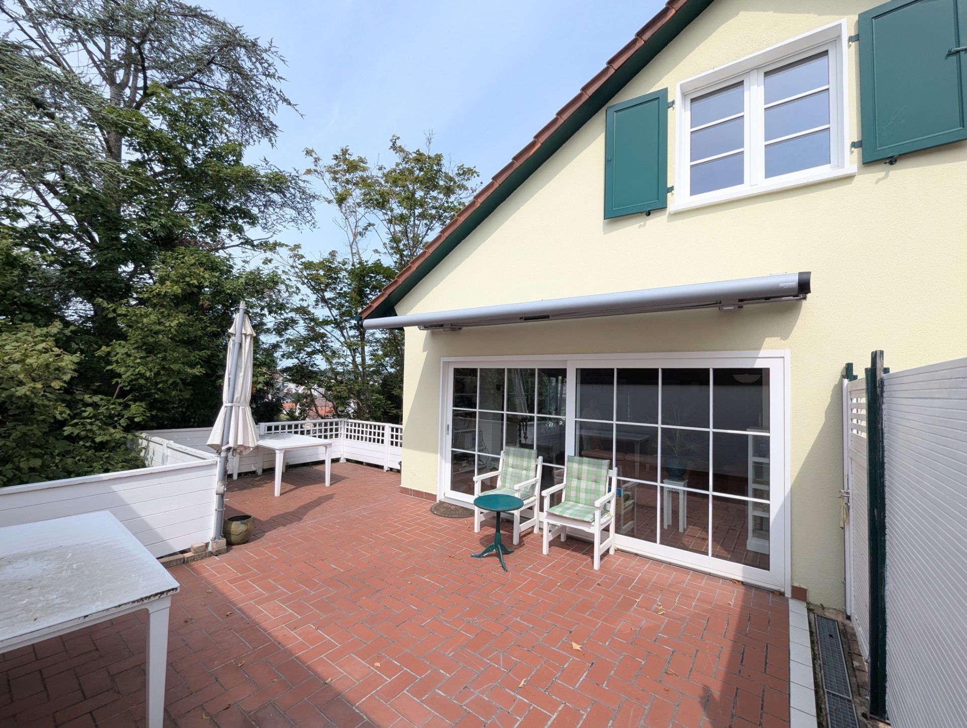 Große Terrasse mit Sitzmöbeln, Tisch, Sonnenschirm und Blick auf Bäume, vor gelbem Haus mit grünen Fensterläden.