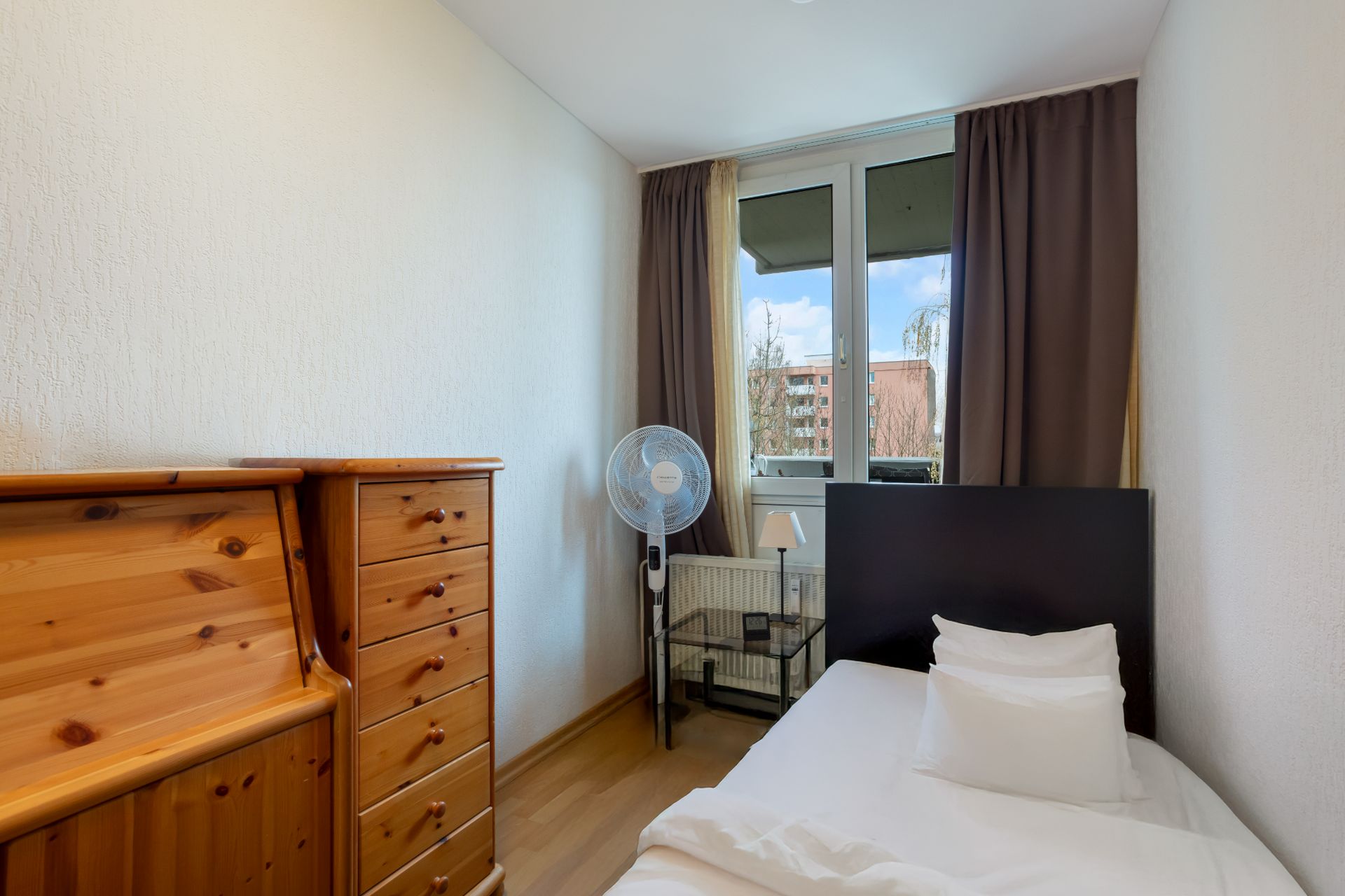 Kleines Schlafzimmer mit Einzelbett, Holzbett, Ventilator und Fenster mit Vorhängen.