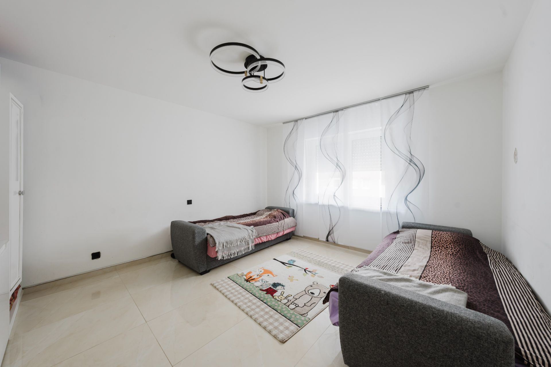 Helles Zimmer mit zwei grauen Einzelbetten, großem Fenster und Teppich mit Tiermotiven.