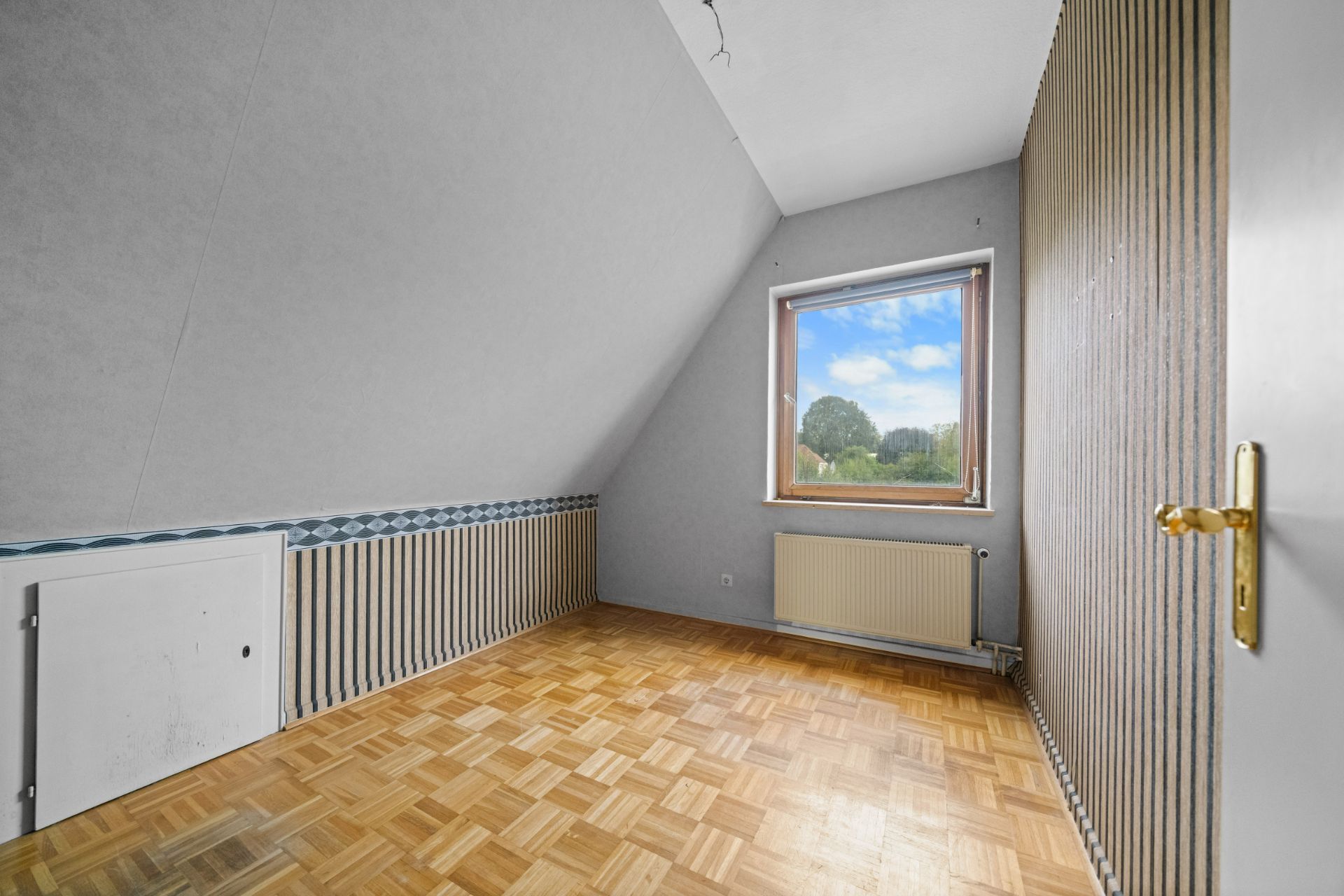 Leeres Zimmer mit Parkettboden, großem Fenster, schräger Decke und Heizkörper unter dem Fenster.