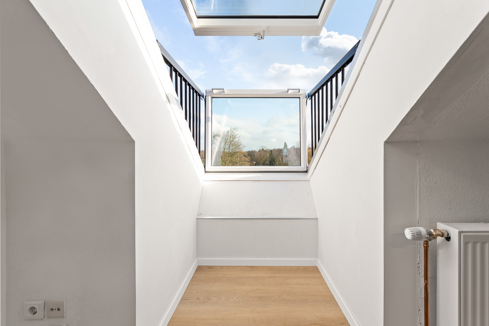 Heller Raum mit Holzfußboden, weißen Wänden und offenem Dachfenster mit Ausblick.