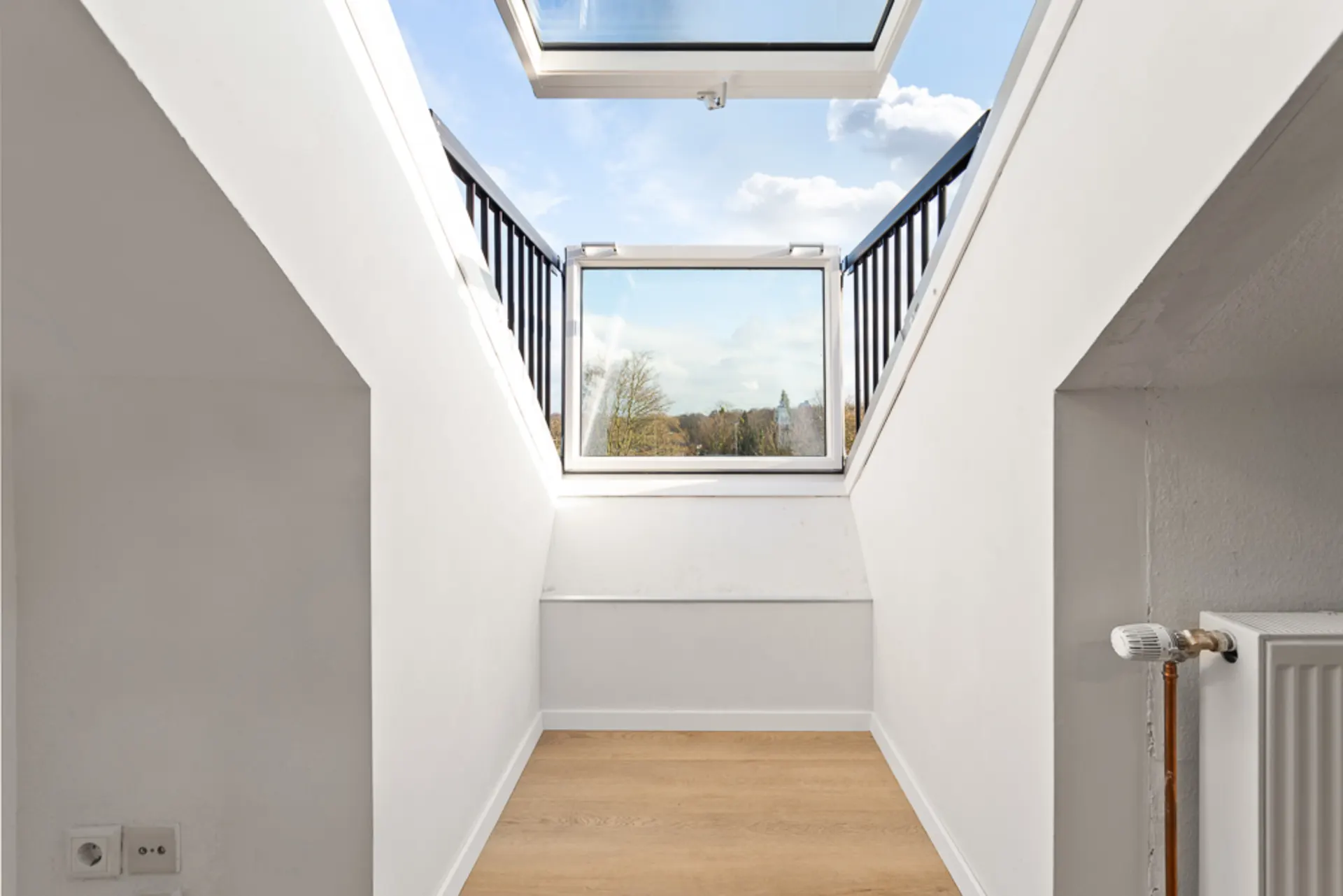 Heller Raum mit Holzfußboden, weißen Wänden und offenem Dachfenster mit Ausblick.